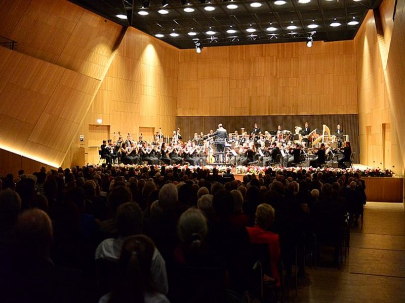 Das Bild zeigt einen vollbesetzten Konzertsaal mit Holzvertäfelung, im Hintergund ist ein großes Orchester auf einer Bühne zu sehen.