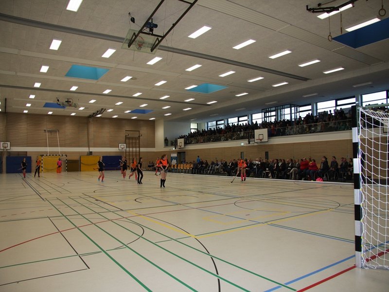 Das Bild zeigt junge Frauen bei einem Hockeyspiel in einer Sporthalle mit gefüllten Zuschauerrängen.