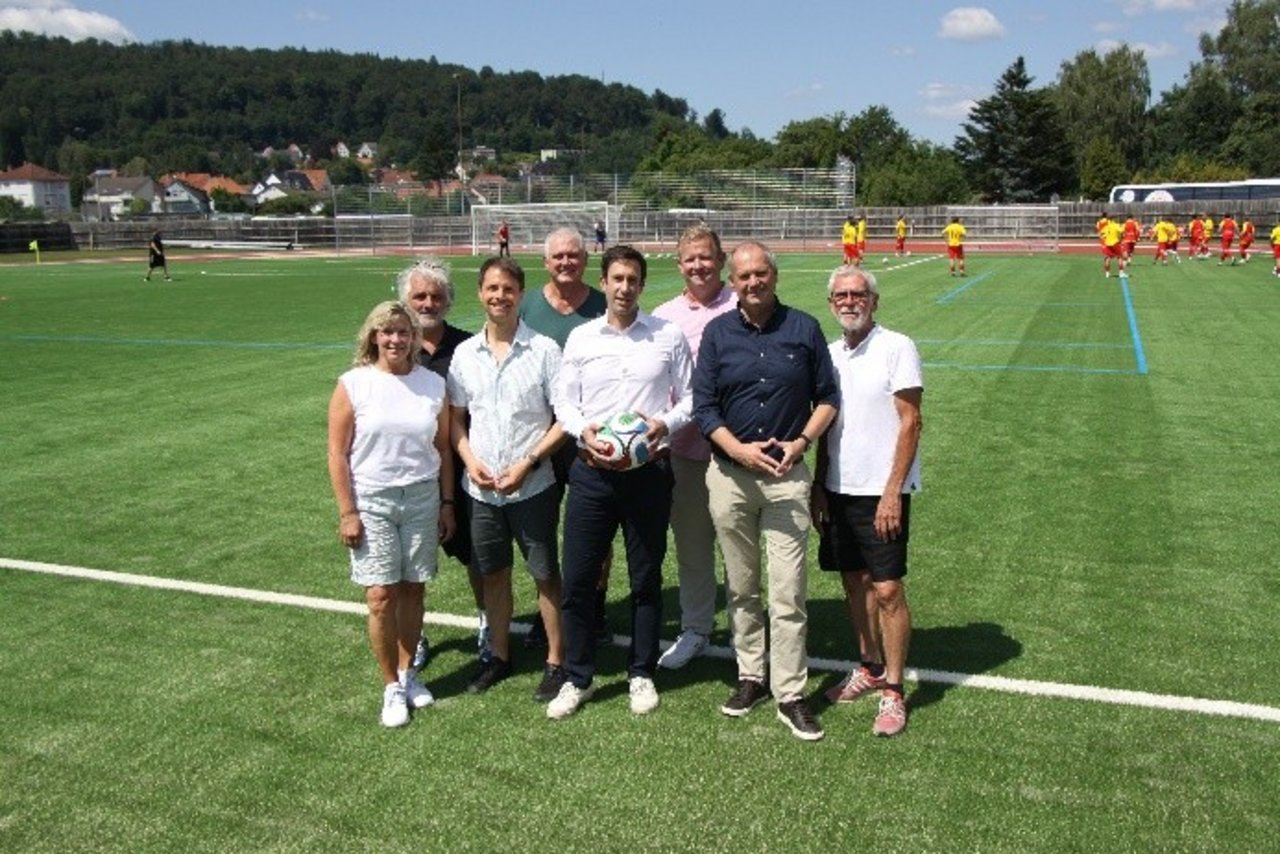Quelle: Stadt Hann. Münden Das Bild zeigt acht Personen bei der Einweihung einer Sportanlage. Im Hintergrund wird Fußball gespielt.