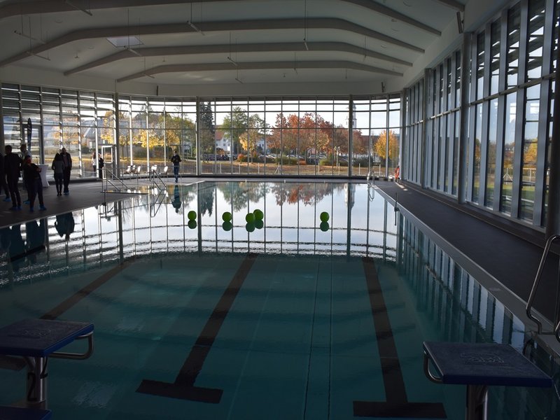 Das Bild zeigt ein Schwimmbecken in einem Hallenbad, im Hintergrund eröffnet eine Fensterfront den Blick nach draußen.