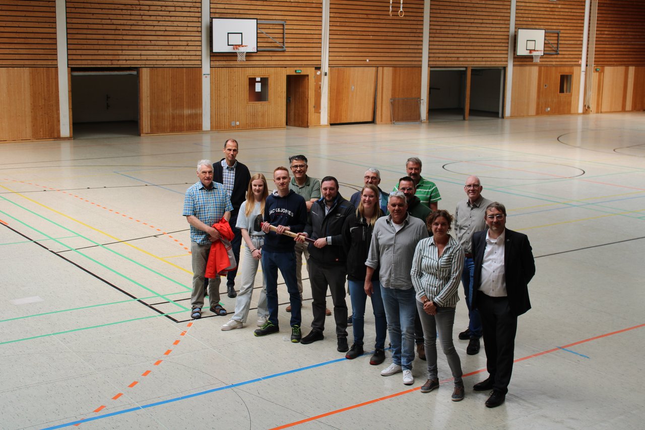 Auf dem Bild sind eine Gruppe Menschen zu sehen, die in der Sporthalle stehen und symbolisch einen Hammer halten. 