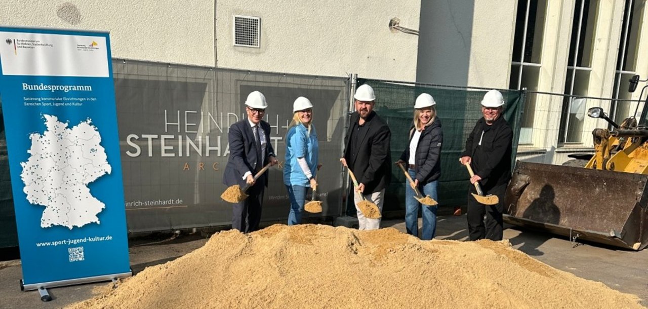 Das Bild zeigt einen Spatenstich. Mehrere Personen mit Baustellenhelm und Schaufel stehen vor einem Sandhaufen und blicken in die Kamera.