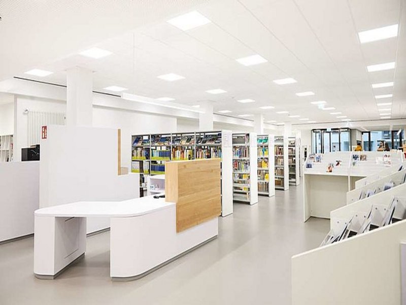 Das Bild zeigt einen vorwiegend weißen Büchereiraum mit Regalen und Tresen.