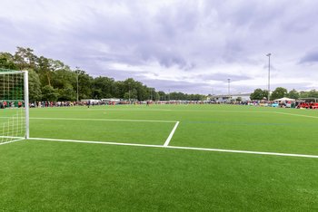 Das Bild zeigt einen Fußballplatz, wo ein Spiel ausgetragen wird. Zuschauer stehen um das Spielfeld herum.