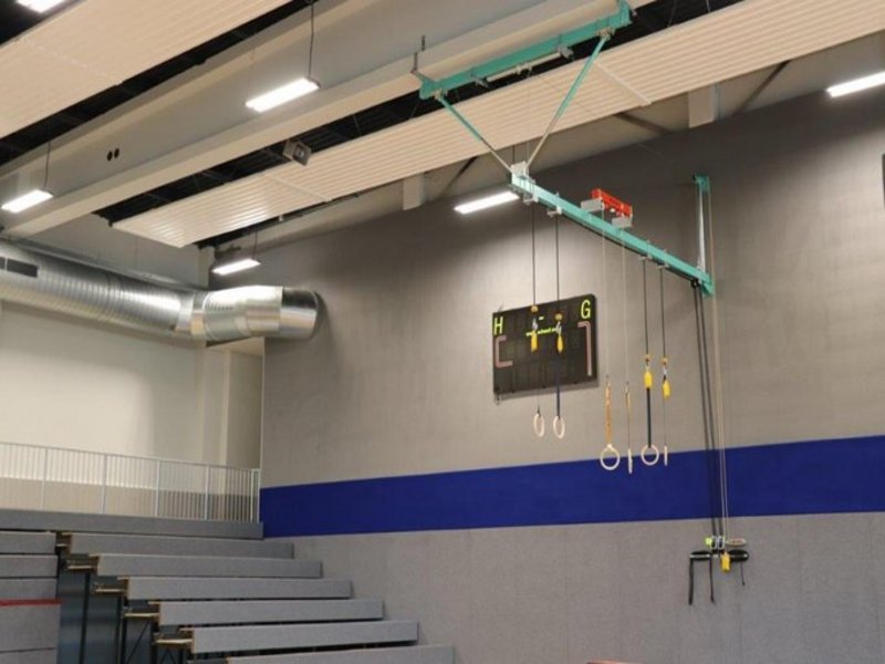 Das Bild zeigt eine Tribüne in einer Sporthalle, außerdem sind Ringe von der Decke hängend und eine elektronische Anzeigetafel an der Wand zu sehen.