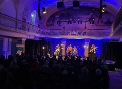 Quelle: Theater Altes Hallenbad Auf diesem Bild sieht man eine Gruppe auf einer Bühne mit Instrumenten die vor eiinem Publikum spielen.