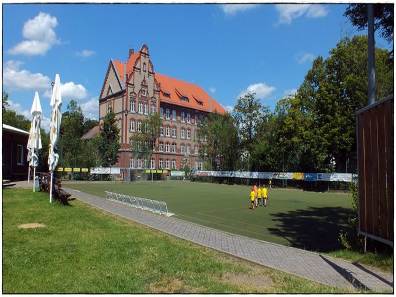 Das Bild zeigt einen Rasensportplatz, im Hintergrund ist ein historisch anmutendes Gebäude zu sehen, einige Kinder betreten gerade den Sportplatz.