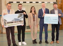 Quelle: Verwaltungsgemeinschaft Schwarzenfeld Man sieht vier Männer und eine Frau, die zwei Spendenschecks hoch halten und vor einem Tor in einer Sporthalle stehen.
