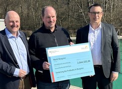 Bildquelle: BMWSB/ Patrick Schneider Das Bild zeigt drei Personen, die vor sich einen überdimensionierten Scheck halten. Im Hintergrund ist ein Schwimmbecken zu sehen.