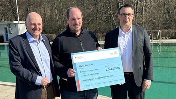 Bildquelle: BMWSB/ Patrick Schneider Das Bild zeigt drei Personen, die vor sich einen überdimensionierten Scheck halten. Im Hintergrund ist ein Schwimmbecken zu sehen.