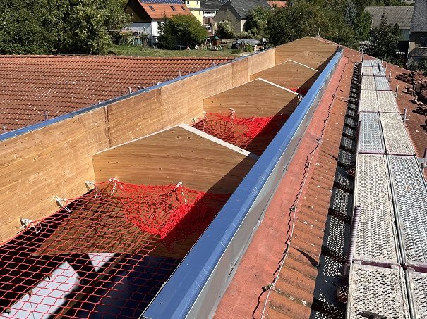 Gemeinde Mohlsdorf-Teichwolramsdorf Bauverwaltung Das Bild zeigt den Baufortschritt am Dach mit einer Holzkonstruktion und roten Sicherheitsnetzen