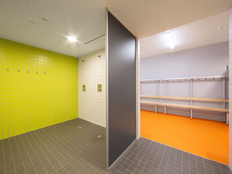 Das Bild zeigt einen Sanitärbereich mit Duschen und den Übergang zum Umkleidebereich mit auffälligen Farbakzenten in gelb und orange.