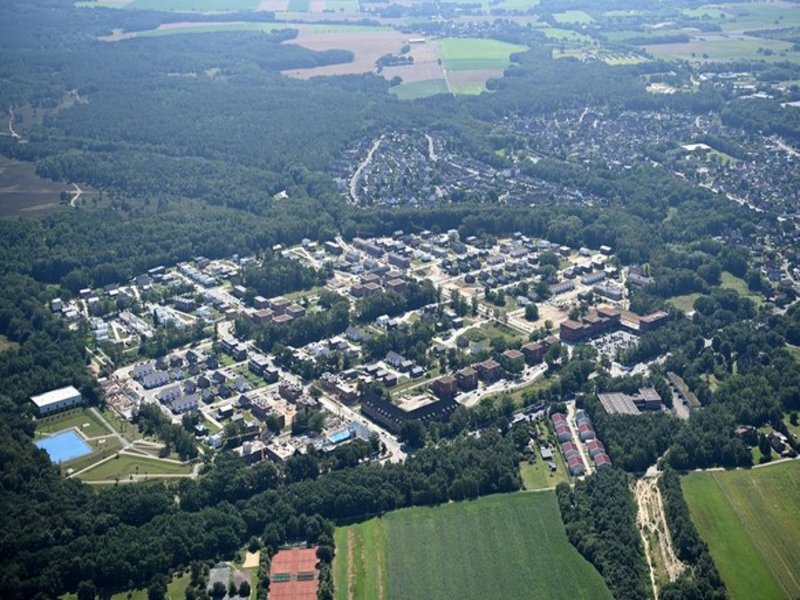 Das Bild zeigt eine Luftaufnahme von einem Gebiet mit mehreren Häusern und einer Sportanlage, umgeben von Bäumen.