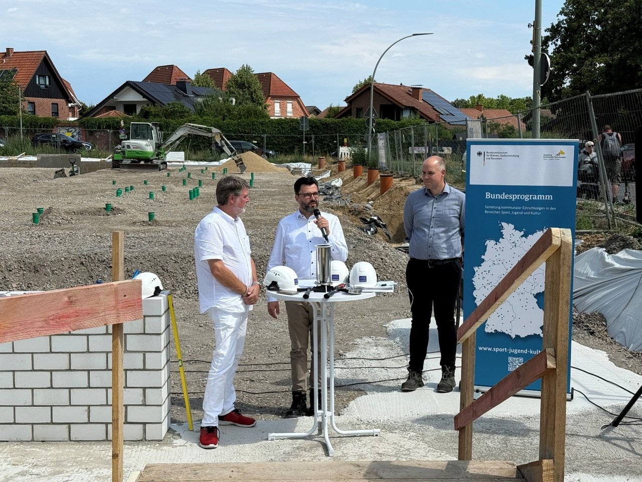 Quelle: Stadt Werne Das Bild zeigt drei Personen vor einer Baustelle. Eine Person spricht in ein Mikrofon zum Publikum.