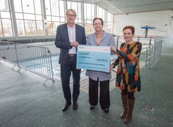 Copyright: Sascha Engst/ Bundesstadt Bonn Man sieht zwei Frauen und einen Mann stehen in einem leeren Hallenbad und halten einen Förderscheck in die Luft.