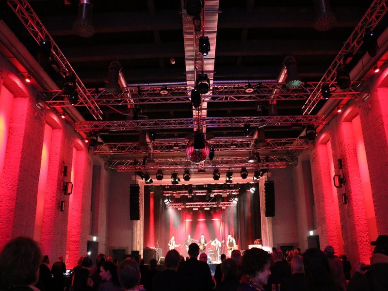 Das Bild zeigt eine Konzertsituation in einem rot beleuchteten Saal mit einer Band auf der Bühne.