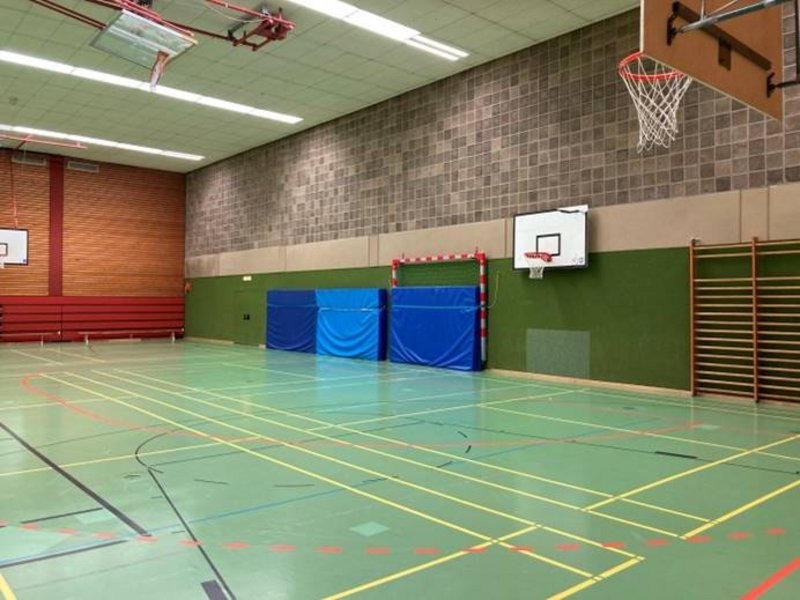 Man sieht das Innere einer Sportturnhalle mit zwei Basketballkörben, drei Weichmatratzen und grünem Boden.