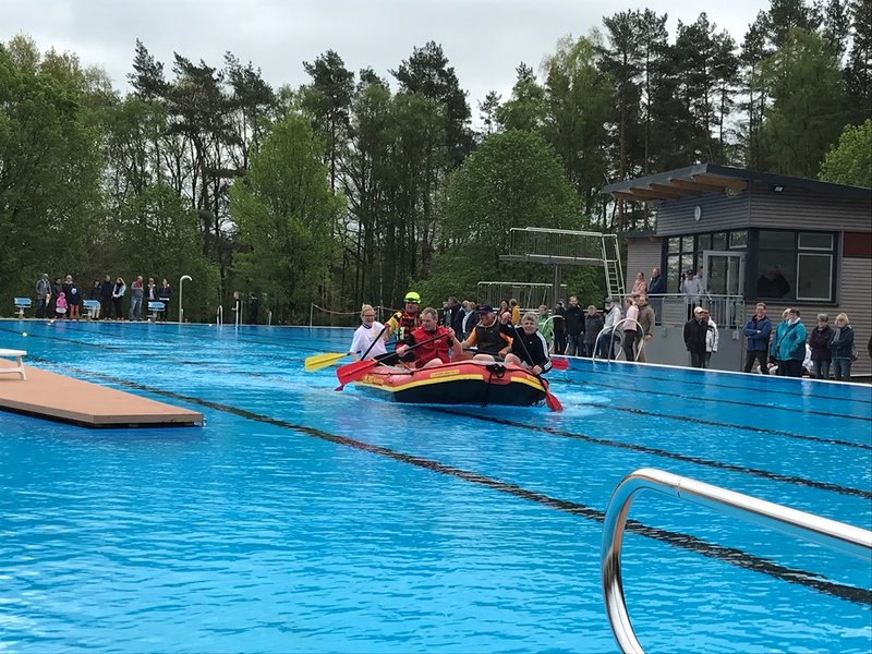 Das Bild zeigt mehrere Personen in einem Schlauchboot, die in einem Schwimmbecken umgeben von Menschen paddeln.