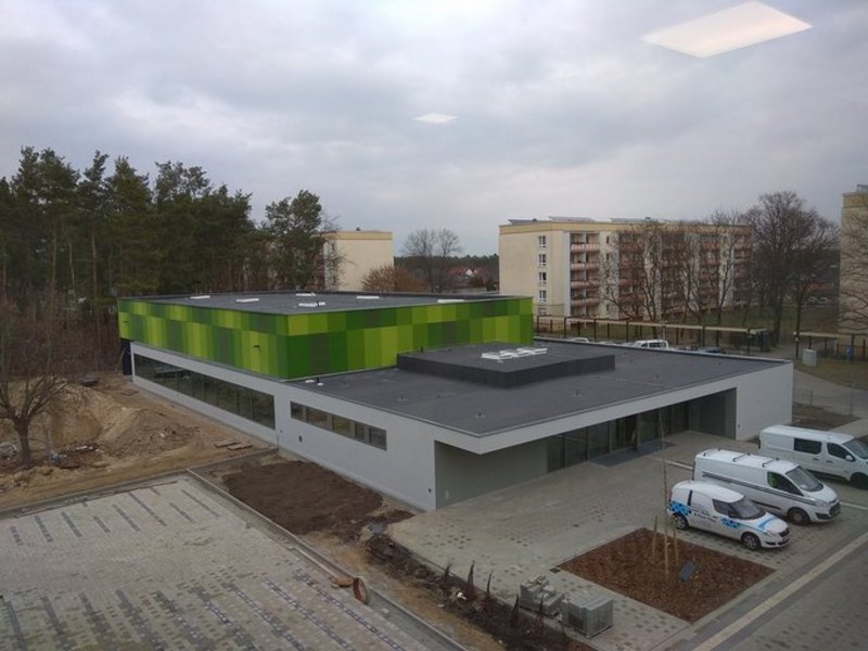 Das Bild zeigt ein modernes graues Gebäude mit Flachdach und grünen Farbakzenten, am Bildrand sind Parkflächen und Autos zu sehen.