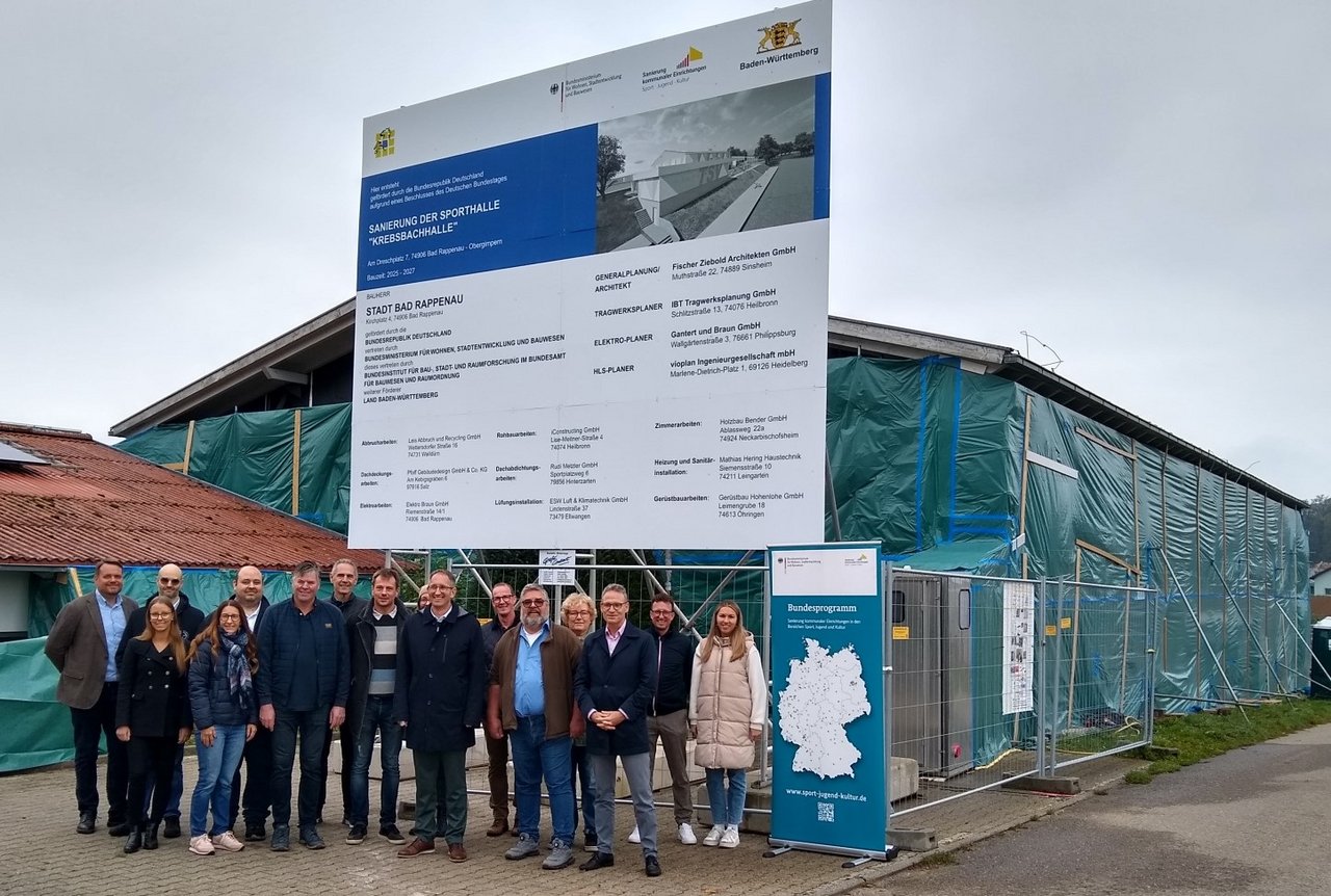 Das Bild zeigt eine Gruppe von Menschen, die vor einer Baustelle mit Bautafel stehen.