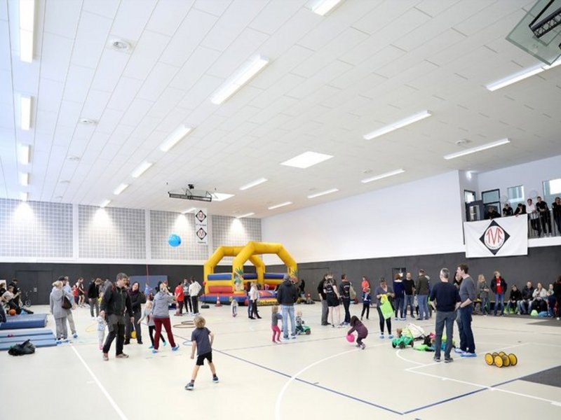 Das Bild zeigt eine Sporthalle, in der Kinder an verschiedenen Stationen spielen und Erwachsene sich in Gruppen unterhalten.