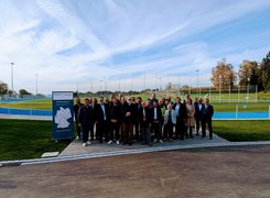 Das Bild zeigt eine Gruppe von Menschen, die vor einer Sportanlage mit Rasen und Flutlichtanlage, die für ein Bild posiert.