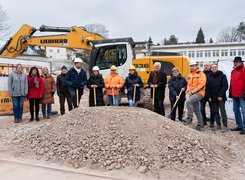 Foto: Gemeinde Heinersreuth Das Bild zeigt eine Reihe von Menschen mit einem Spaten in der Hand. Sie stehen vor einem Bagger. Vor ihnen ist ein Sandhaufen zu sehen.
