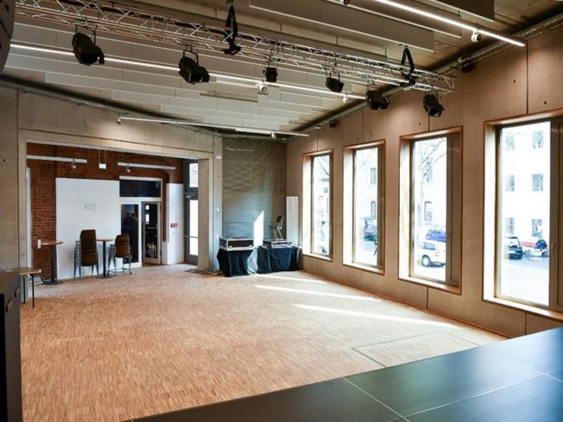 Das Bild zeigt einen leeren Raum mit mehreren Fenstern und Beleuchtungstechnik an der Decke, im Hintergrund sind gestapelte Stühle und Technikkoffer auf Tischen zu sehen.