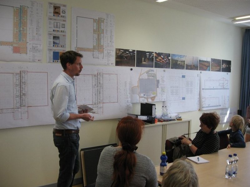 Das Bild zeigt einen Mann, der an einem Bauplan an der Wand einigen Menschen um sich etwas erläutert.