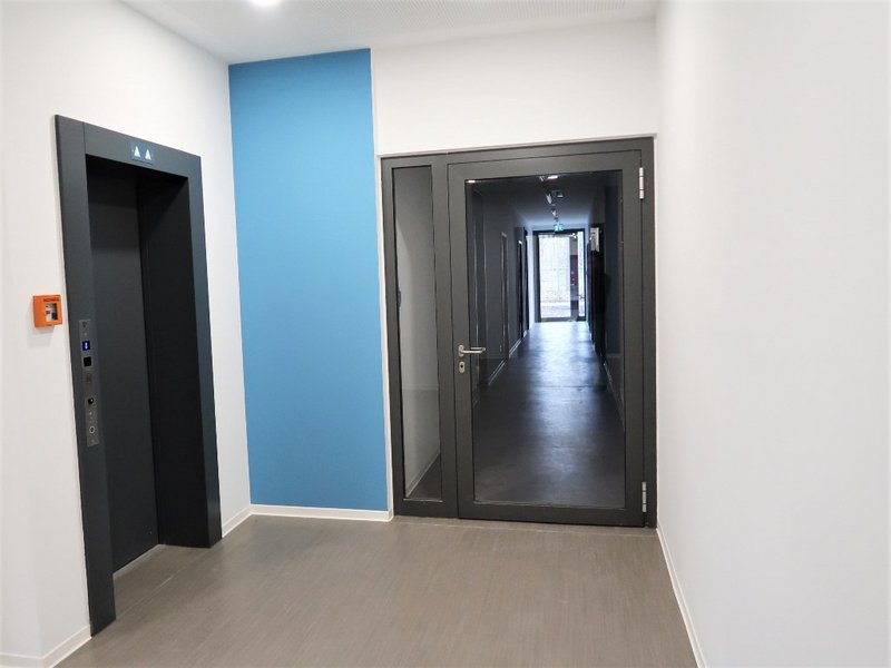 Das Bild zeigt den Zugang zu einem Flur und eine Aufzugtür, dazwischen ist ein blaues Wandelement zu sehen.