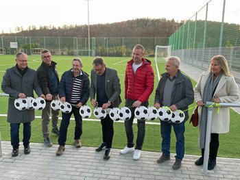 Sieben Personen stehen vor einem Fußballplatz. Sie halten ein gelbes Seil gespannt, auf dem schwarz-weiße Fußbälle aufgehängt sind. Der Mann in der Mitte ist dabei die Schnur durchzuschneiden.