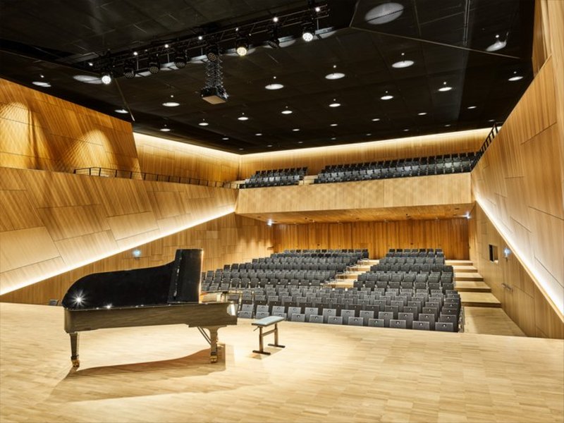 Das Bild zeigt einen leeren Konzertsaal mit Holzvertäfelung, heller Beleuchtung und Balkon, im Vordergrund ist eine Bühne mit Flügel und Hocker zu sehen.