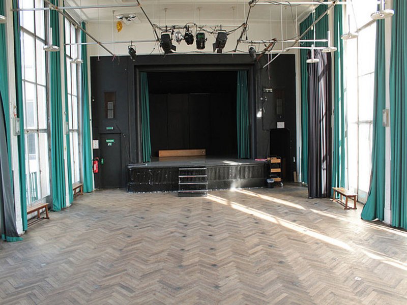 Das Bild zeigt einen Saal mit Bühne, Beleuchtung und türkisen Vorhängen an den Fenstern links und rechts.