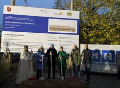 Man sieht sechs Personen mit Spaten die vor einem Bauschild zur Einfachsporthalle Kleinkahl stehen.