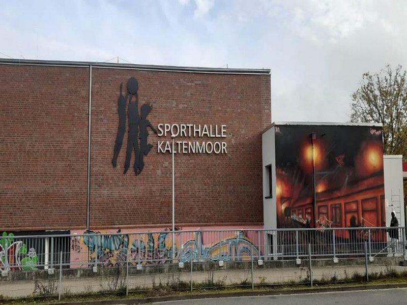Das Bild zeigt eine Backsteinmauer mit dem Schriftzug Sporthalle Kaltenmoor und einen Anbau mit einem Graffiti.