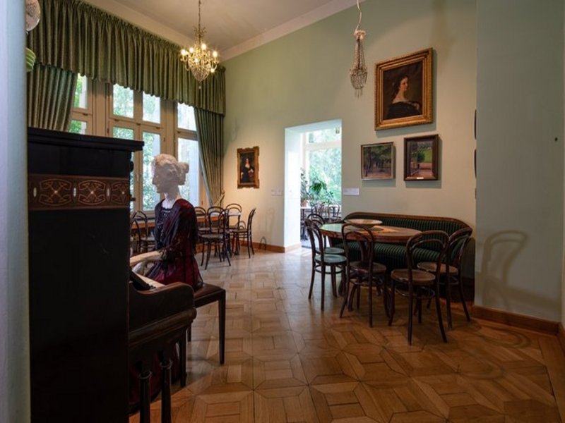 Das Bild zeigt einen Innenraum mit Tischen und Stühlen, es ist eine Puppe an einem Klavier sitzend zu sehen.