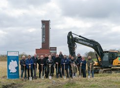 Das Bild zeigt eine Menschen, die symbolisch Spaten in der Händen halten. Im Hintergrund ist ein Bagger und ein hoher Turm zu sehen.