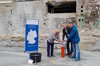 Zwei Männer und eine Frau stehen vor einer Wand. Sie befüllen ein orangenes Rohr mit Zeitungen. Das symbolisiert die Grundsteinlegung an ihrem Projekt. Links von den Personen steht ein Aufsteller und ein Baueimer.