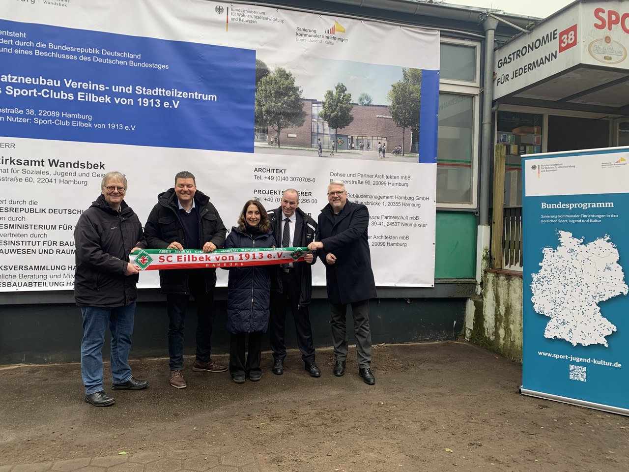Quelle: Freie und Hansestadt Hamburg, Bezirksamt Wandsbek Fünf Personen stehen vor einem Plakat, das die Planungen für den Neubau des Vereinsgebäudes ankündigt. Lachend präsentieren sie einen Vereinsschal mit dem Schriftzug des SC Eilbek von 1913 e.V.