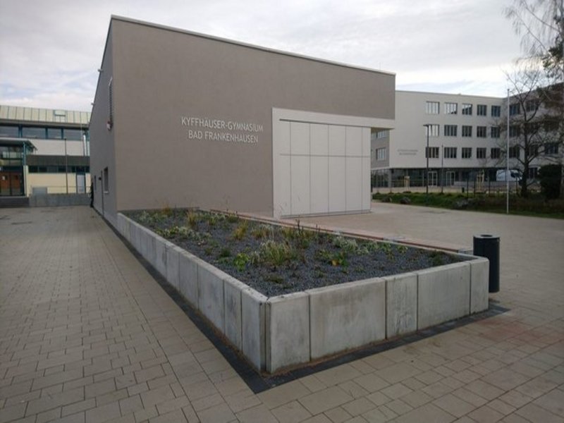 Das Bild zeigt die Hauswand eines Gebäudes mit Flachdach, die den Schriftzug Kyffhäuser Gymnasium Bad Frankenhausen trägt, im Hintergrund ist ein größeres Gebäude in ähnlichem Stil zu sehen, im Vordergrund eine Bepflanzung.