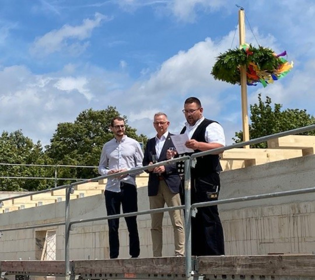 Förderverein Freibad Remlingen Das Bild zeigt drei Männer auf einer Tribüne. Im Hintergrund ist ein Baum mit Kranz zu sehen.