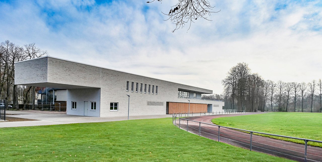 Quelle: Architektur Büro Müntinga Schimmelpfeng Das Bild zeigt einen Rasenplatz mit Laufbahn und einem neuen Gebäude.