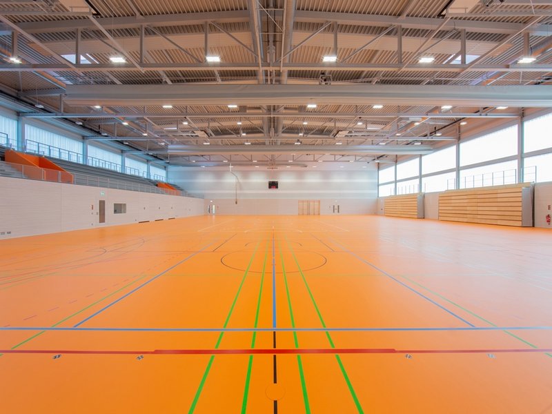 Das Bild zeigt eine Sporthalle mit orangenem Boden und heller Wand- und Dachverkleidung.