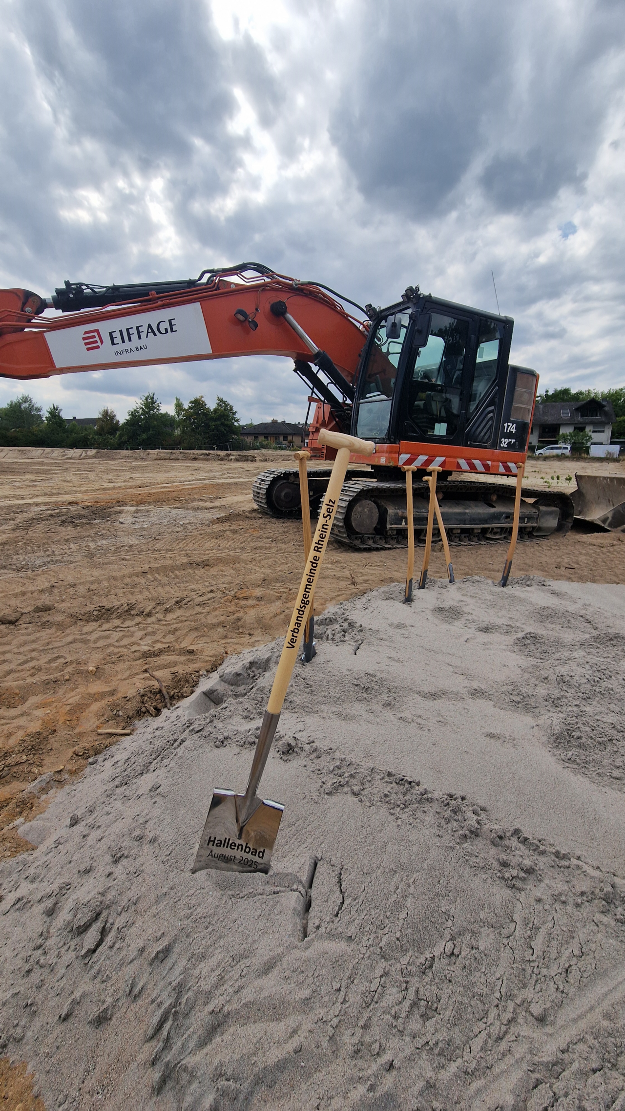 Quelle: Verbandsgemeinde Rhein-Selz Das Bild zeigt einen Erdhügel, in dem ein Spaten steckt, im Hintergrund sind Baumaschinen zu sehen.