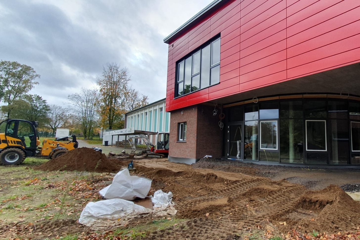 Das Bild zeigt die neue Sporthalle mit roter Fassade, großen Fenstern und Baustelle mit Erdarbeiten und Baumaschine im Vordergrund.