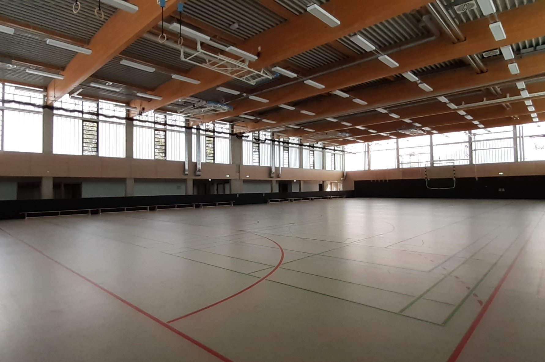 Stadt Winsen (Luhe) Das Bild zeigt die Innenansicht einer sanierten Sporthalle mit Holzbalkendecke, großen Fenstern und markiertem Spielfeldboden.