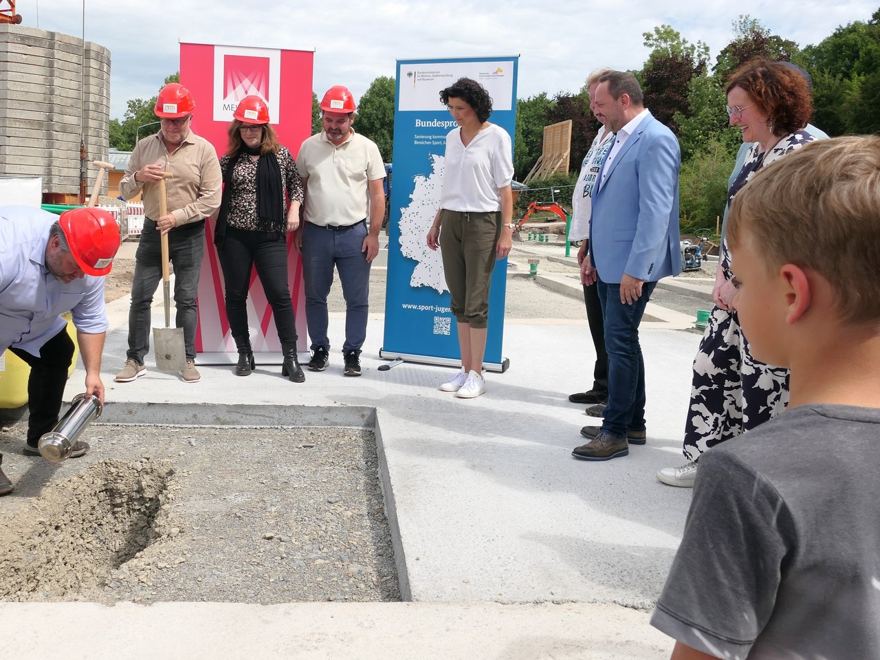 Das Bild zeigt sieben Personen, die um eine Grube herum stehen. Eine Person legt eine Zeitkapsel in das Fundament um die Grundsteinlegung zu symbolisieren..