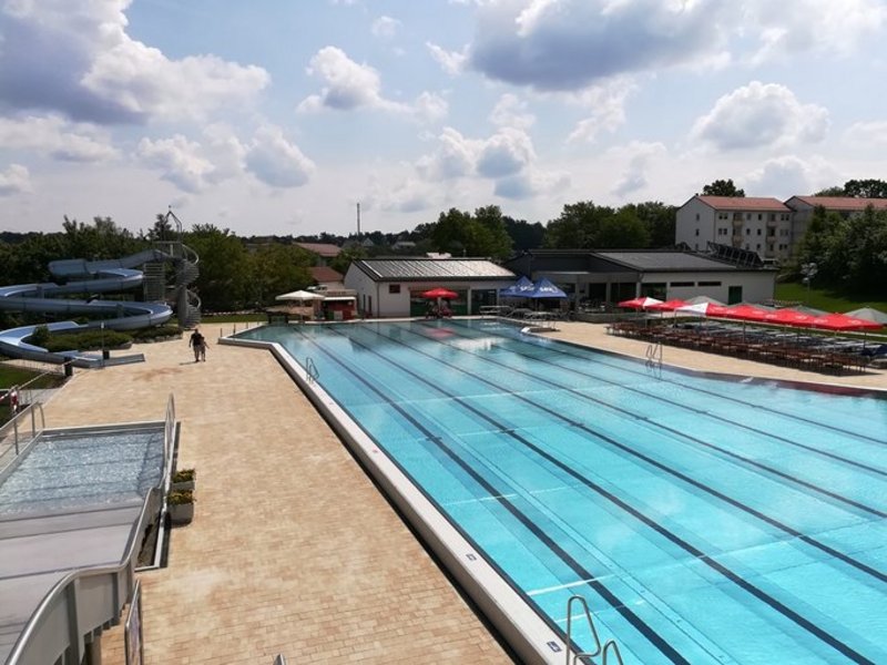 Das Bild zeigt ein menschenleeres Schwimmbecken mit heller Pflasterung außen herum, im Hintergrund eine Rutsche und ein Gebäude zu sehen.
