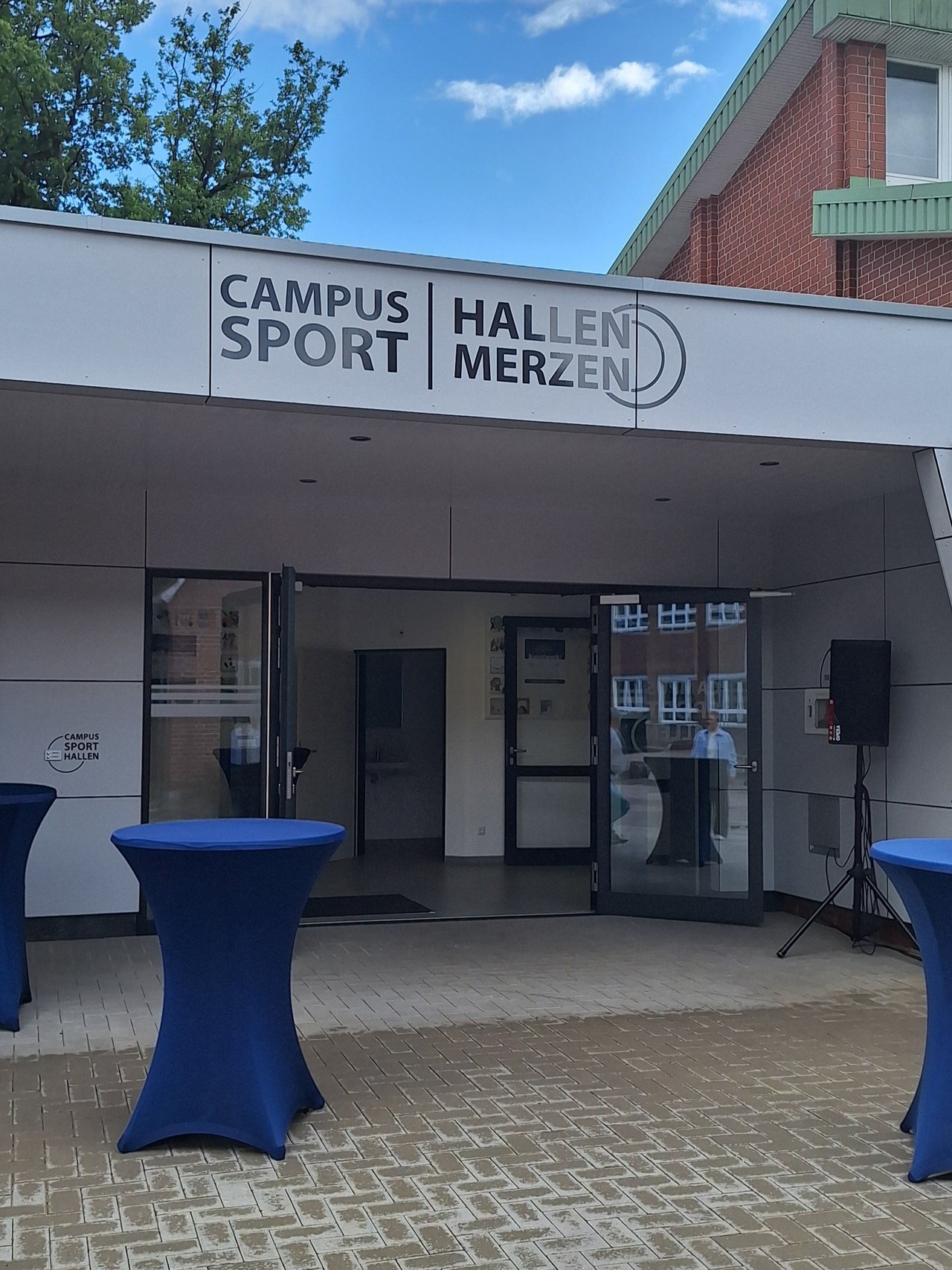 Das Bild zeigt den Eingang einer Sporthalle von außen. Davor stehen Stehtische mit blauen Hussen.