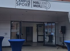Das Bild zeigt den Eingang einer Sporthalle von außen. Davor stehen Stehtische mit blauen Hussen.